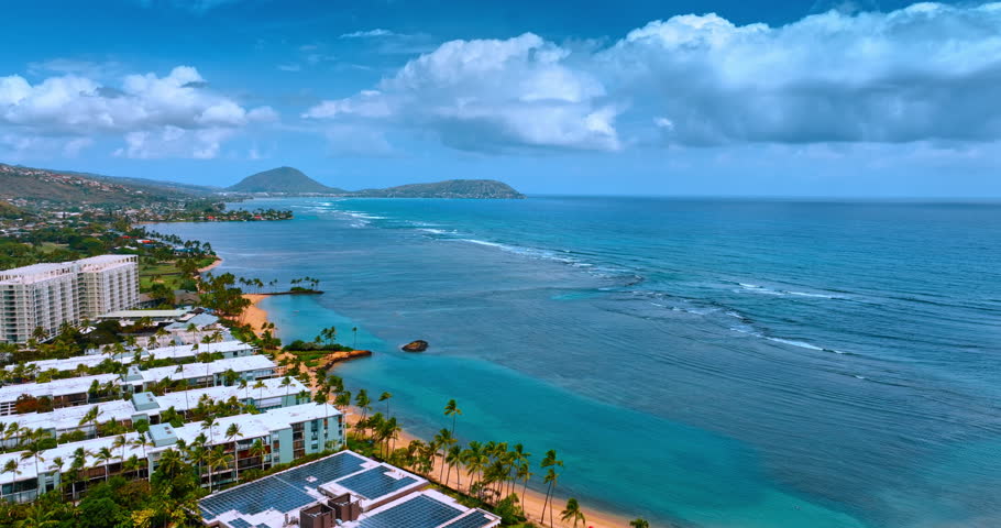 Built-up shore of Honolulu, Hawaii with sandy beach and palm trees. Pacific Ocean waves roll slowly to the coast. Clouds hang in sky over the waterscape.