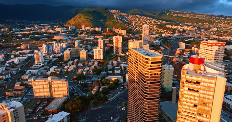Bright orange light reflecting on the facades of the buildings. Dark dramatic clouds roll on the mountains at backdrop. Honolulu, Hawaii, USA.