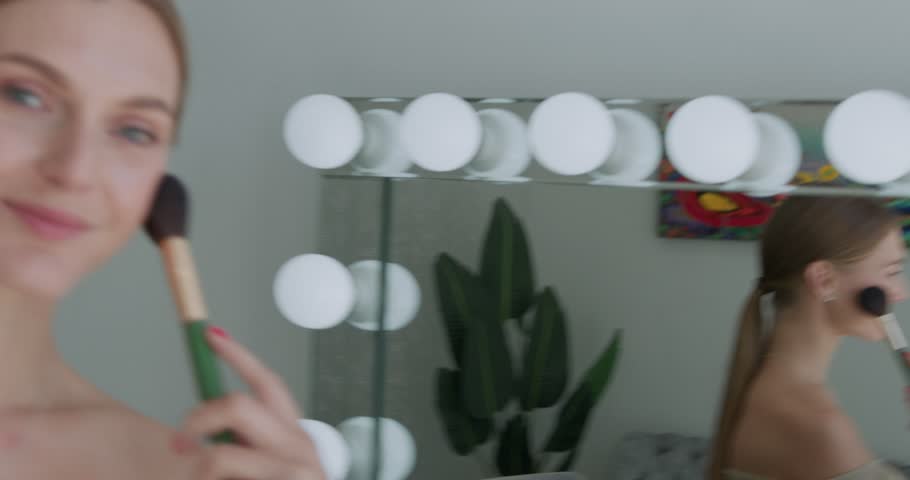 A girl with a makeup brush poses for the camera, confidently smiling in front of a mirror.