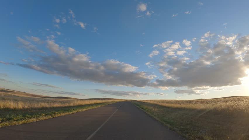 POV from the of vehicle while it drive along a rural prairie road towards a setting sun
