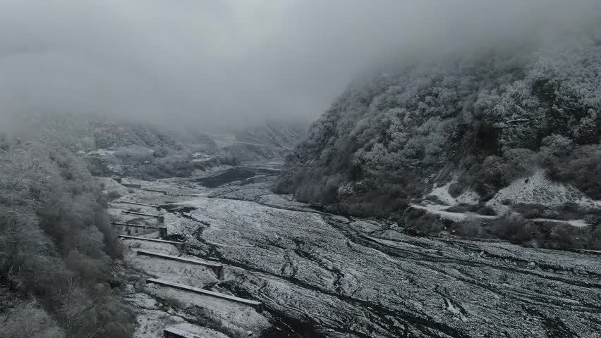 A drone flies towards ethereal, snow-dusted hills shrouded in thick fog. A mysterious and serene winter atmosphere.