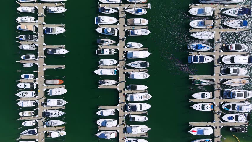 Top view of marina with many docks and white boats. Boats are lined up in rows on green water. Neat layout shows organized space for sailing and relaxing near coast.