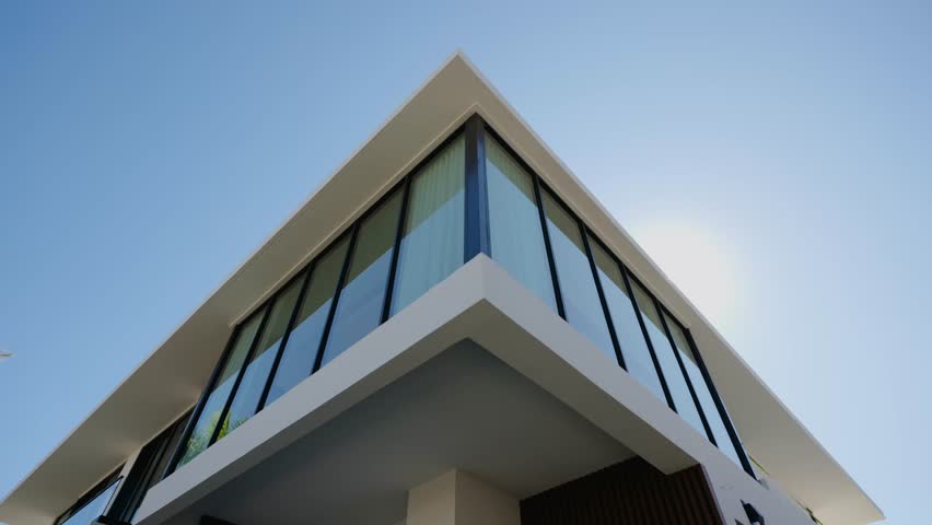 Corner of modern building with large glass windows and white roof stands under clear blue sky. Sharp lines and bright light create clean, open, and stylish look.