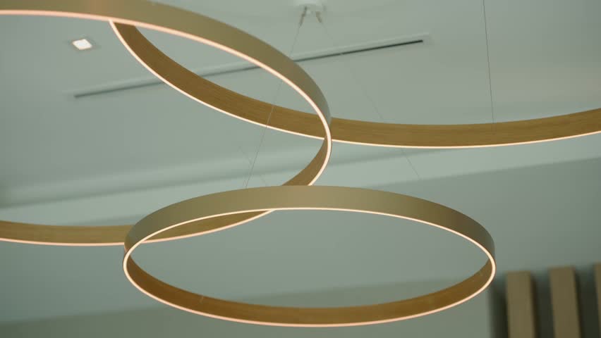 Circular ceiling lights hang in soft white space. Rings glow gently from inside edges, floating on thin wires. Design looks clean, modern, and artistic.