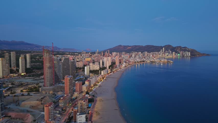 Aerial Curving Beach At Blue Hour Showing Long Shoreline, Gentle Waves And Illuminated Promenade Ideal