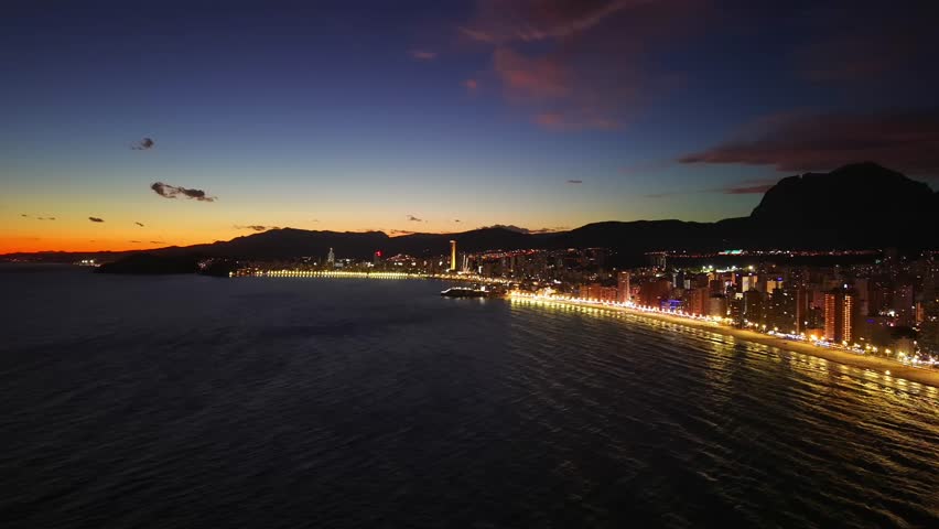 Aerial Illuminated Promenade Along Sandy Beach At Dusk With Shimmering Sea Reflections And Mountain Backdrop,