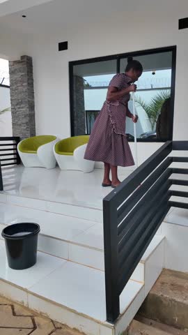 Lilongwe Malawi Africa, 01.12.2025. Video. African female worker with mop and bucket cleaning an outdoor patio and steps of a modern apartment in Lilongwe Malawi 