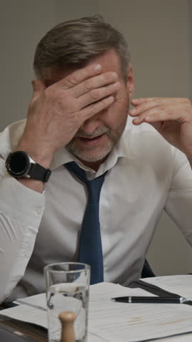 Medium vertical shot of frustrated, annoyed Caucasian male executive or barrister, wearing white shirt and tie, examining business contract or case paperwork, rubbing face and drinking water