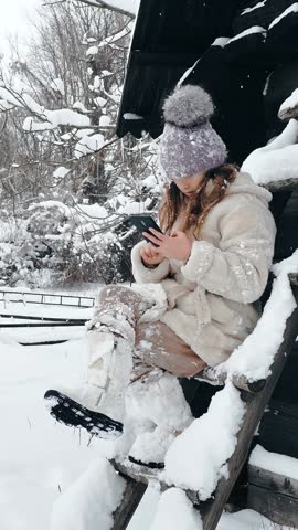 Snowy winter fun. cutie girl, dressed in warm winter clothes, fully covered with snowflakes, is sitting on bench and using smartphone, outdoors, during snowfall, near old wooden house. winter day.