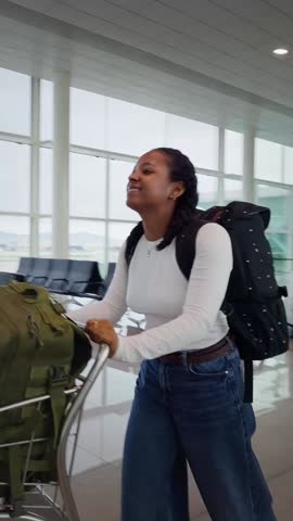 Vertical. African millennial woman pushing a luggage trolley through a bright airport terminal, walking past the glass walkway toward the gates. Travel mood shows movement, anticipation and calm