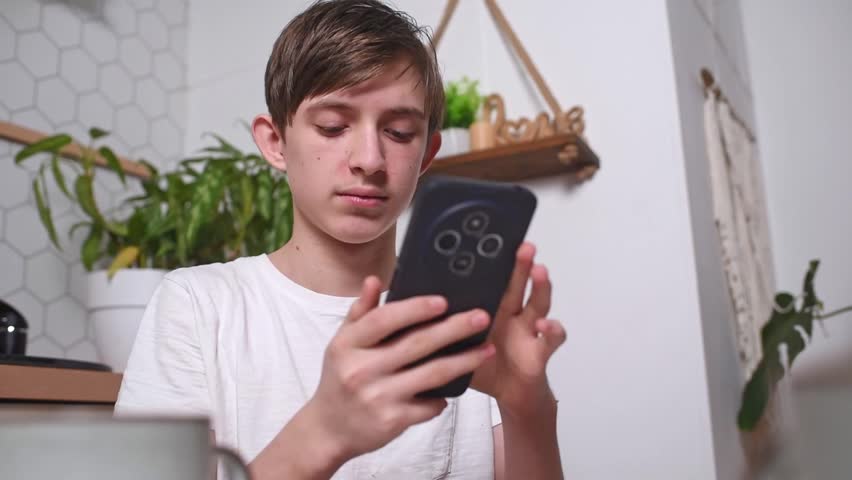 A teenage boy holds a smartphone in his hands, surfing the Internet during breakfast in the kitchen.