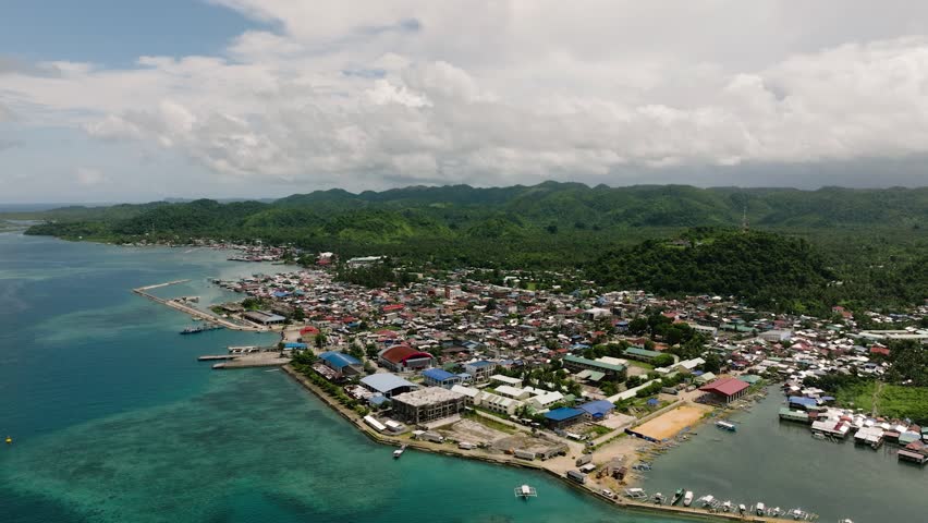 A coastal town with colorful buildings stretching inland against a backdrop of green hills. Del Carmen. Siargao, Philippines.