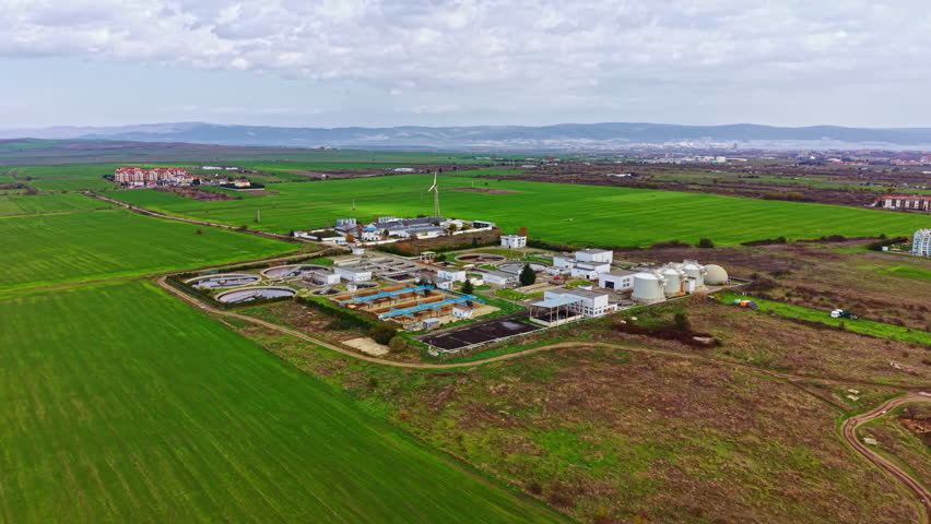 In a lush green setting, a state-of-the-art wastewater treatment facility stands prominently, surrounded by fields and gentle hills, with the clouds softly floating overhead.