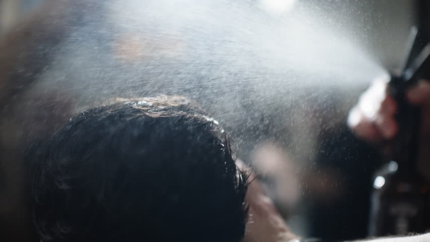 Backlit grooming moment as a stylist applies a fine mist of hairspray to freshly styled hair, captured in close-up with shimmering droplets and soft bokeh in a bustling salon setting.
