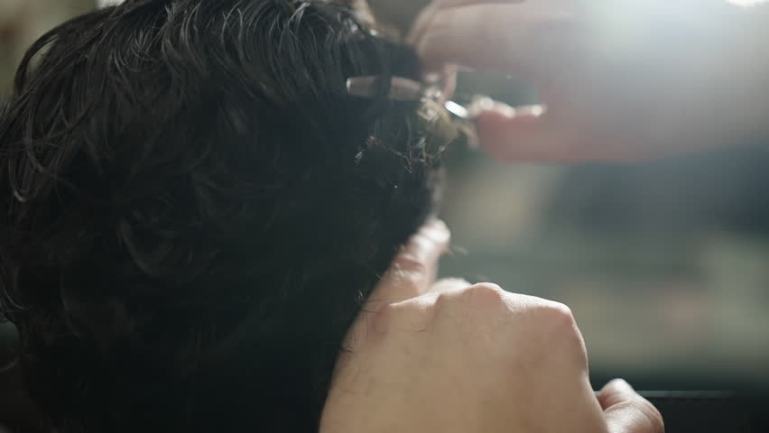 Close-up of a barber cutting wavy dark hair with scissors and comb, carefully trimming around the ear and nape as a client sits in a salon, lit by soft natural light.