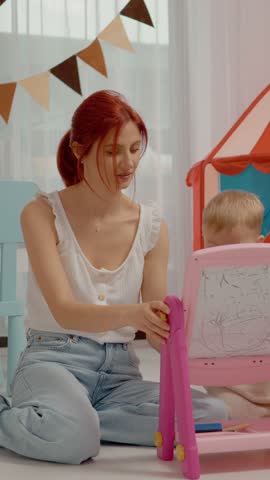 Vertical Screen: A mother spends quality time with her young son as they draw on a blackboard. The playful setting includes toys and a colorful indoor tent for added fun.