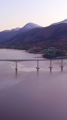A tranquil scene of a long bridge stretching across calm waters with majestic snow-capped mountains in the background at twilight