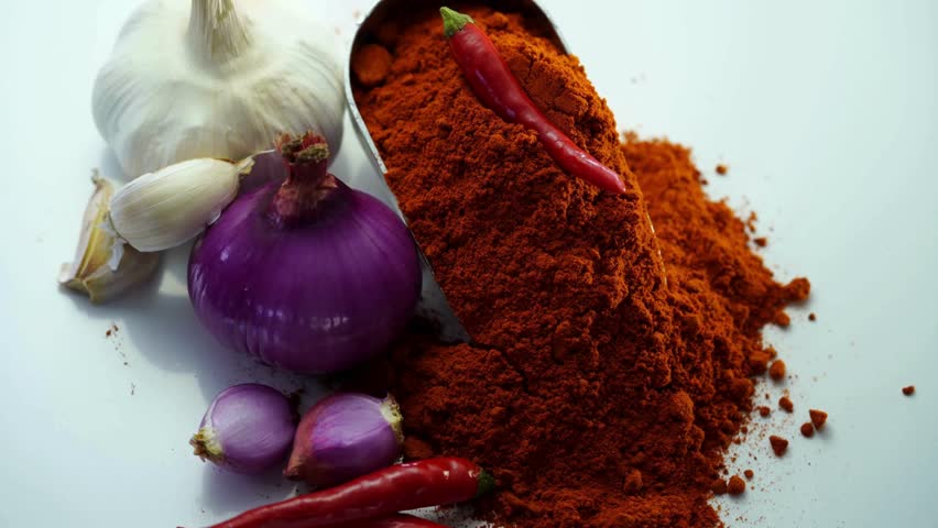 A metal scoop filled with red chili powder resting on a white surface beside garlic and a whole chili pepper
