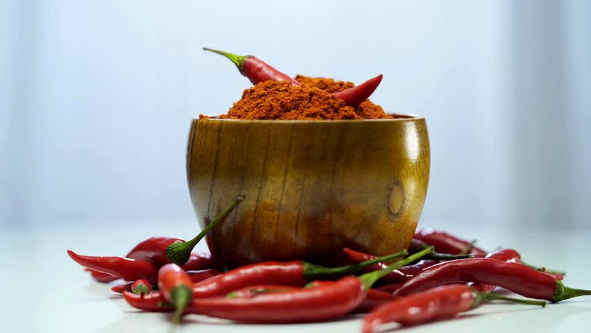 A wooden bowl filled with vibrant red chili powder surrounded by whole red chilies on a bright clean surface