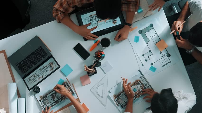 Top view of smart architect engineer writing at blueprint while sitting at meeting table with house model and laptop. Skilled manager working and taking about about building structure. Alimentation.