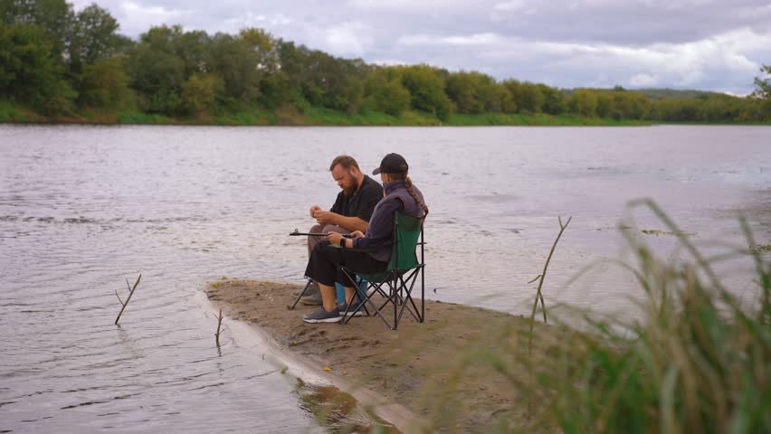 Young couple spending quality time together, sitting on folding chairs by the river and preparing their fishing rods for a day of angling, enjoying a quiet weekend getaway in nature