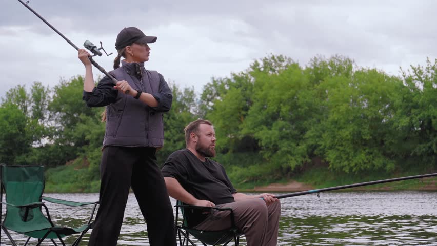Woman standing on the riverbank casting a fishing line while a man sits beside her in a chair, both enjoying a relaxing day of fishing together outdoors in a beautiful natural setting