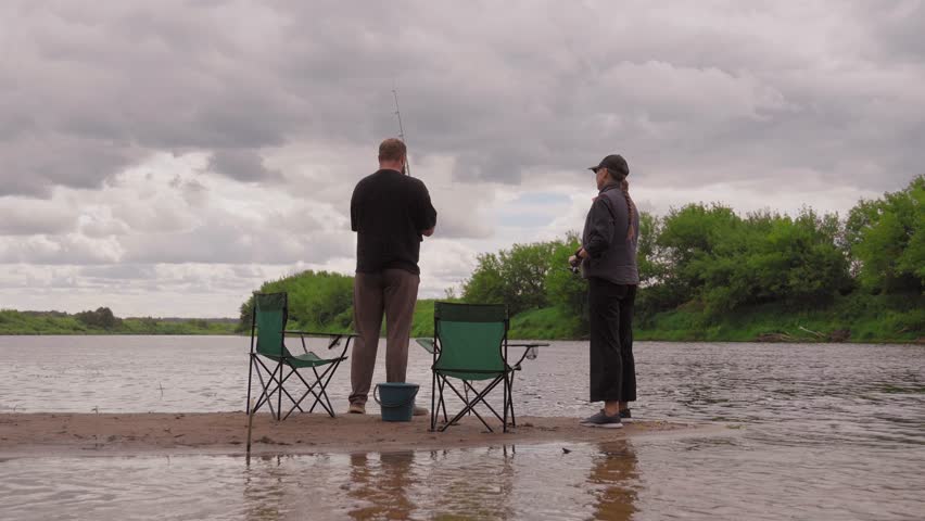 Two people, a man and a woman, fishing with spinning rods from a sandy bank in the middle of a river under a cloudy sky, enjoying their outdoor hobby and spending leisure time together
