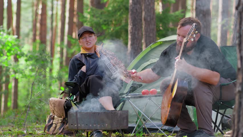 Young couple enjoying a romantic picnic at their campsite in the woods while the bearded man plays the guitar for his smiling girlfriend near the tent and smoking campfire on a sunny day