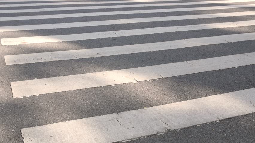 person walking across pedestrian zebra crosswalk with soft sunlight and shadow, man wearing jeans and leather shoes movement across zebra crossing on asphalt road, city life commuting safety