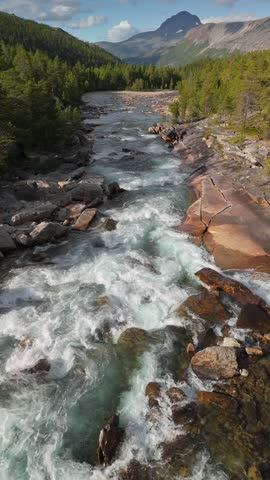 Rushing river flows through rocky landscape in mountain region. Flying over a fast mountain river surrounded by mountains and forests. Aerial shot