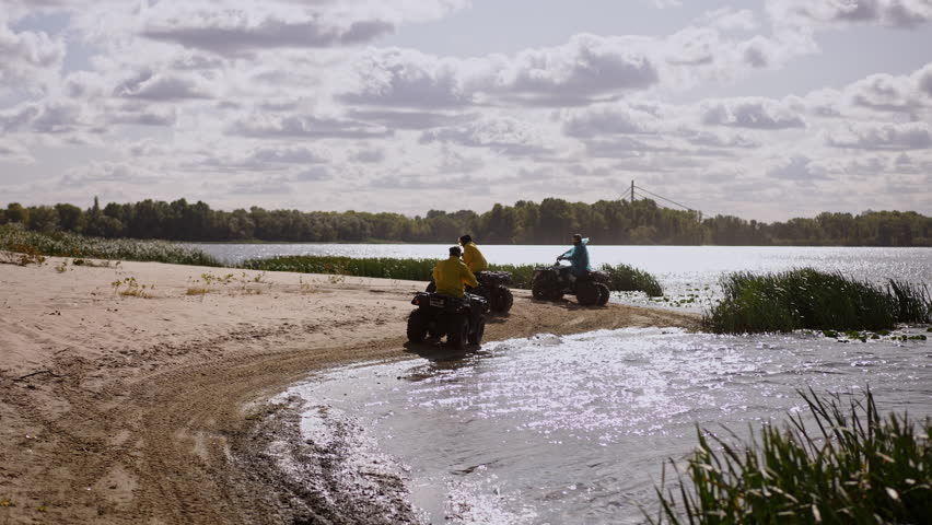 Group quad ride along sandy coast under cloudy sky, convoy of riders in bright jackets crossing shallow tidal pool, water glinting in sunlight, dune grasses