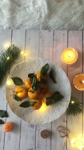 Tangerines lie on a stand on a wooden table. Candles burn nearby, and a garland glows. New Year