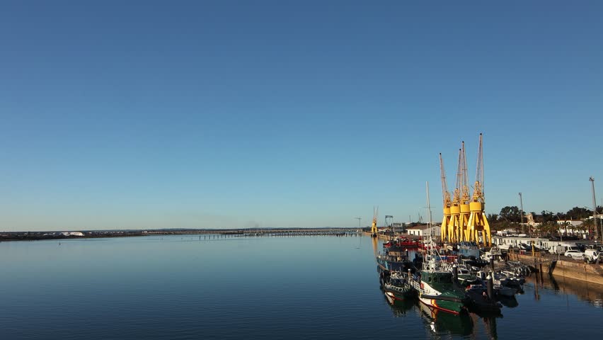 Wide aerial view of the industrial Huelva port, featuring vibrant yellow cranes and docked vessels reflecting on the calm, blue waters of the river or estuary.