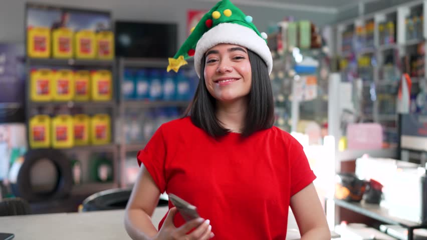 Young latina woman wearing a christmas hat showing a smartphone screen at an auto parts store