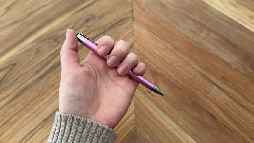 Hand clicks a pink pen repeatedly while holding it over a wooden table. Subtle fidgeting motion representing boredom, restlessness, or distraction. Shot in 4K.