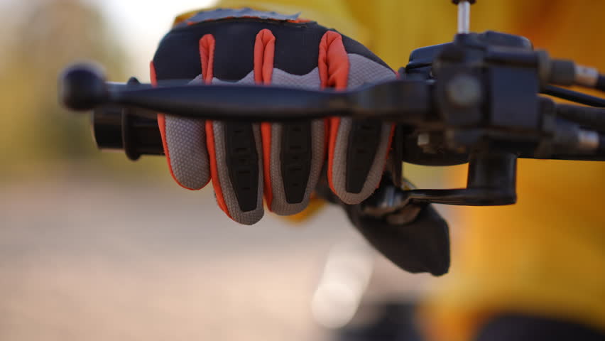 Gloved hand on motorcycle throttle, closeup view of rider adjusting grip and brake lever, orange accented protective glove and yellow jacket sleeve, shallow