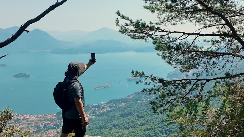 Tourist photographing lake panorama with smartphone from mountain summit. Capturing scenic alpine landscape through phone.