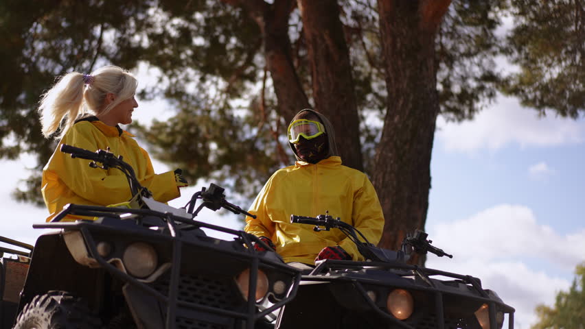 Slow motion. Closeup riders on quad celebrate with high five, two friends in matching yellow jackets and helmets laugh under sunlit pine, dynamic movement, upbeat