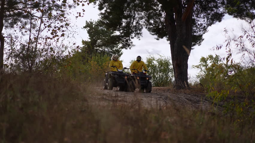 Slow motion. Distant riders on atv trail under tree, two people in yellow jackets pause beside large pine, muted autumn light, grassy foreground, slow contemplative
