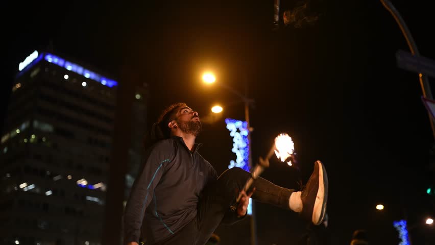 Charismatic male street artist juggling with flaming torches at a city intersection at night. The talented performer entertains with a dangerous fire show