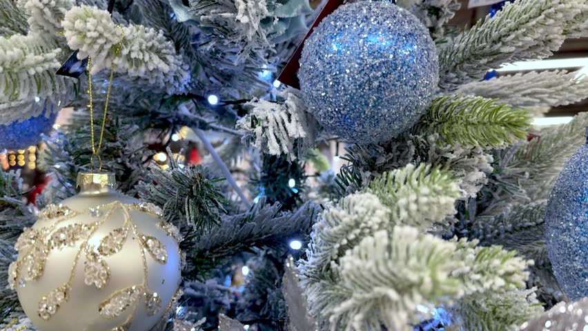 Closeup of snow flocked Christmas tree features elegant silver and blue ornaments. Decorations include sparkling glitter against white branches and blue string lights. New Year 4k footage.