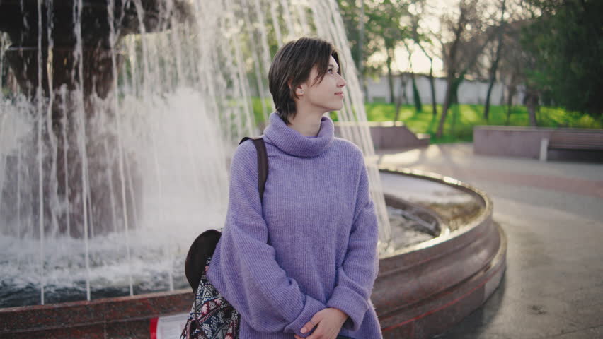 Young woman sits by a fountain in a park during the day, looking towards the sky near trees and benches in the distance on a pleasant afternoon