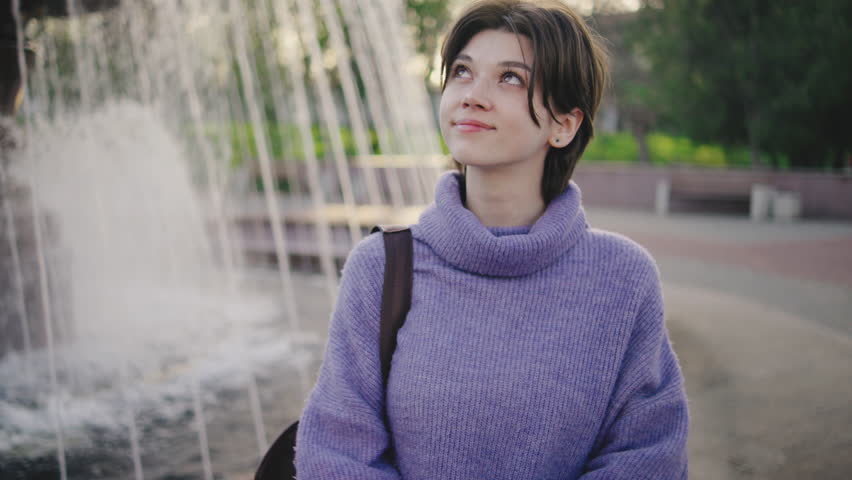 Young woman enjoys time near fountain in park during afternoon hours while looking up with a thoughtful expression and wearing a knitted sweater