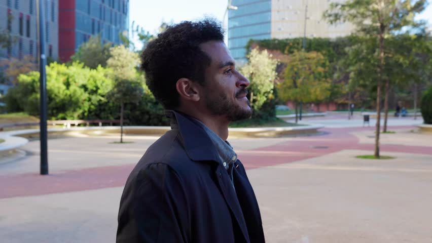 Young handsome businessman wearing a trench coat walking outdoors through a city park during the daytime. Stylish and confident entrepreneur commuting to his office in the financial district