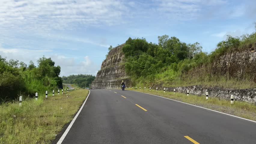 Rural road running through green hills and rocky slopes, peaceful countryside transportation scene in natural landscape.