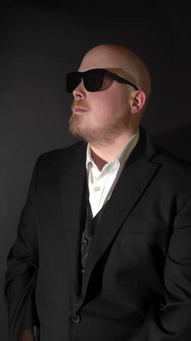 Confident adult man wearing a suit and sunglasses looks directly into the camera and suddenly punches forward against a dark studio background.