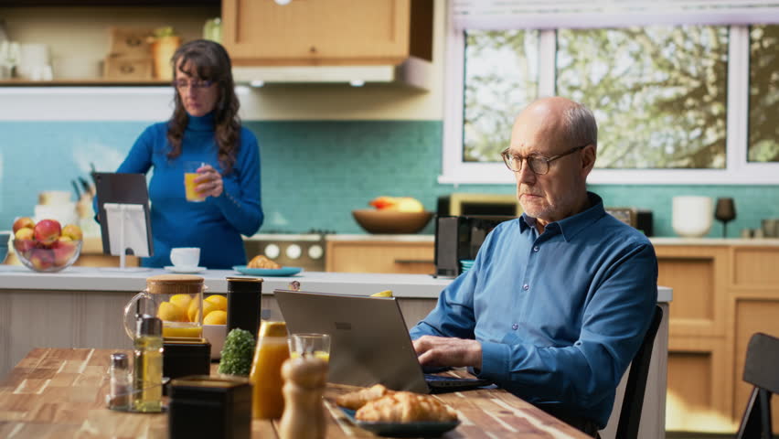 Unhappy distressed old man finding bad disappointing new on laptop, his elderly wife is consoling him and being supportive. Couple serving coffee at the breakfast table. Camera B.