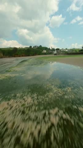 FPV glide over grassy marsh of Brittany bluff overlooking wide beach, soft waves, and bright horizon, vertical