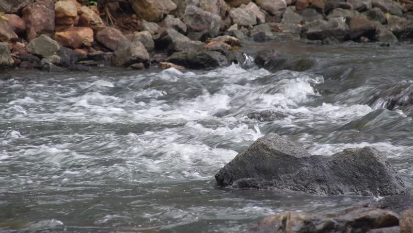 A fast flowing stream with turbulent into white rapids and foam. The rushing water crashes against a steep bank composed of diverse rocks and stones.A natural atmosphere for relaxation on holiday.