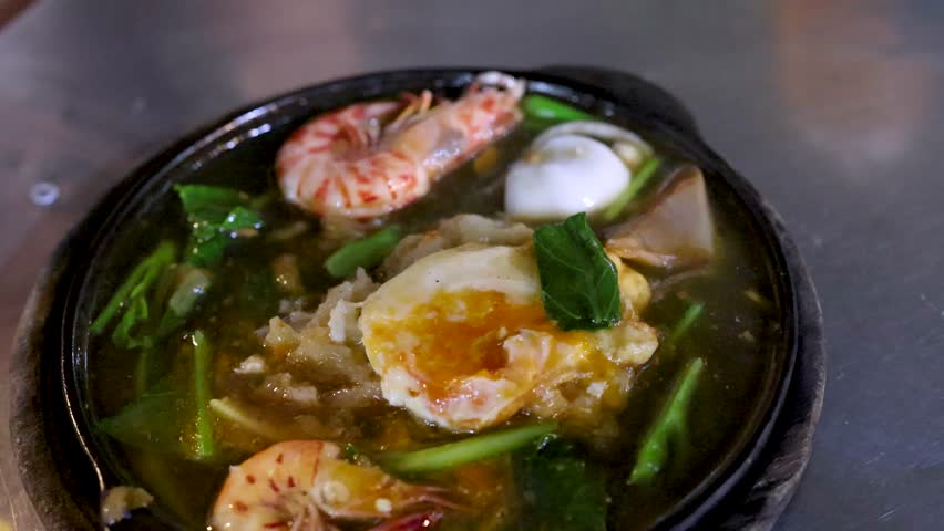 Hand uses chopsticks to mix steaming pad thai noodles, shrimp, vegetables on sizzling hot plate